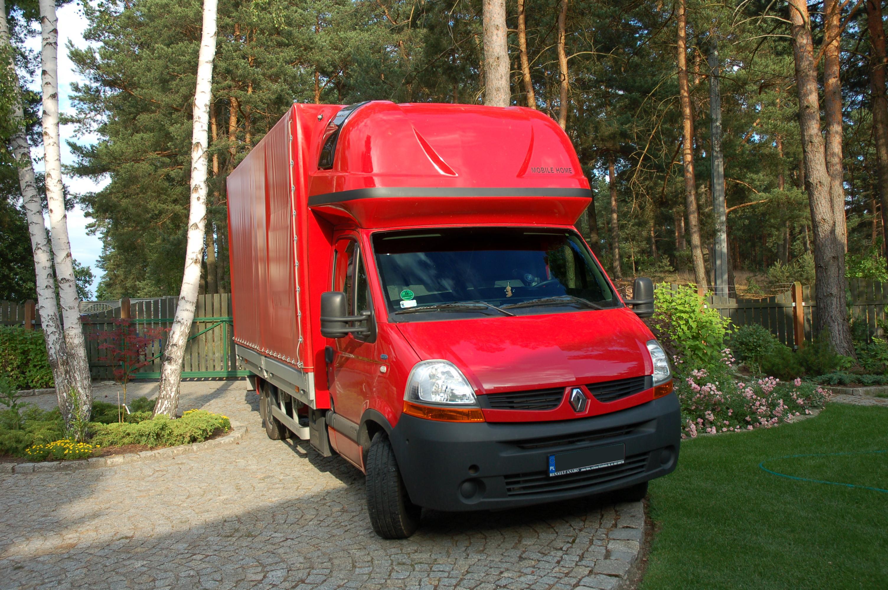 Czerwony samochód dostawczy Renault z plandeką, zaparkowany na brukowanej posesji przed domem, z widocznym napisem 'MOBILE HOME' na nadbudowie kabiny.